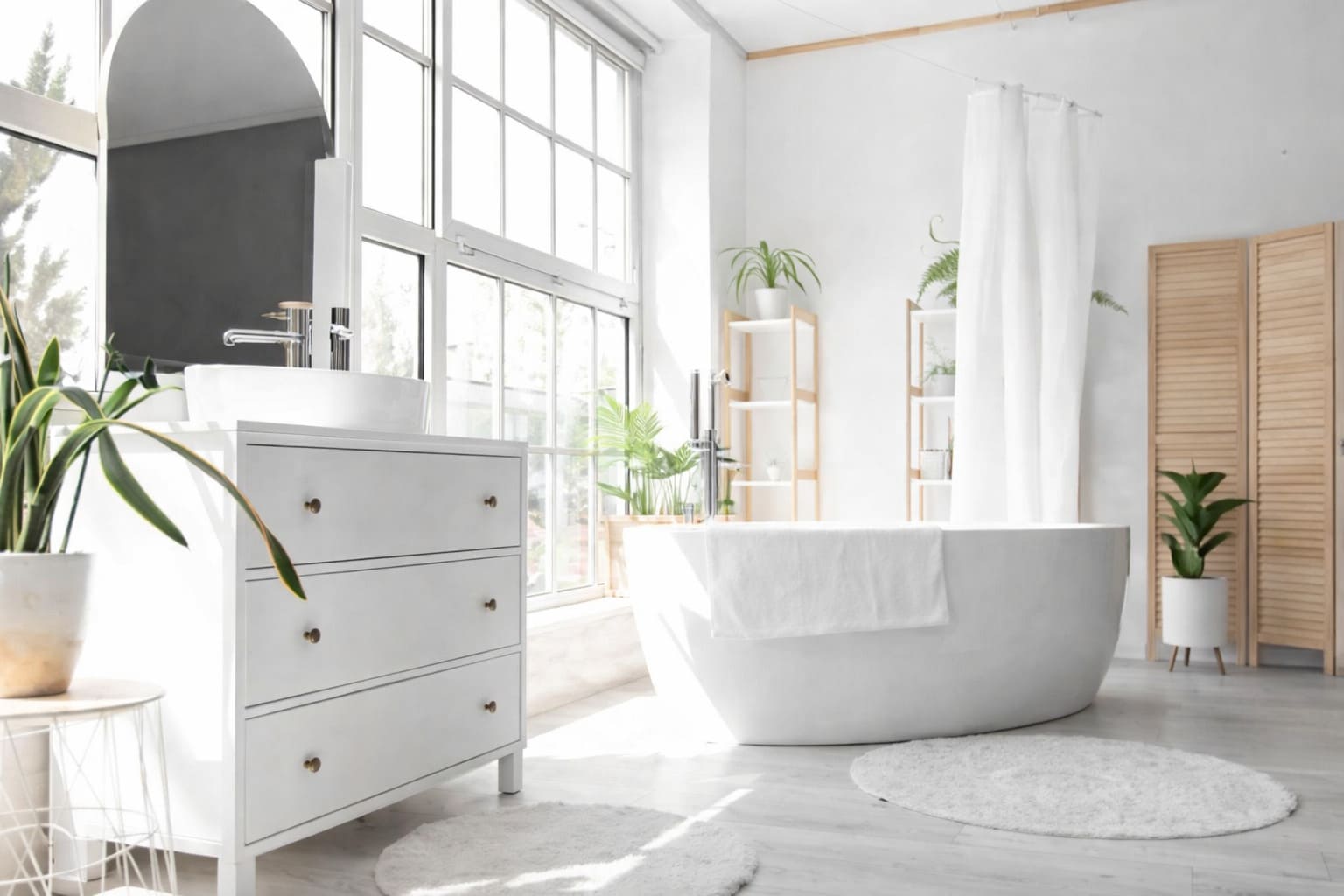 Bright modern bathroom with a freestanding bathtub, white vanity, indoor plants, and large windows.
