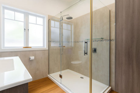 A modern bathroom with a glass shower enclosure, beige wall tiles, and a bright window providing natural light.