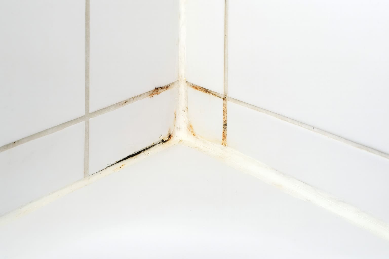 Bathroom tile corner with cracked caulk and stained grout showing mould and water damage.