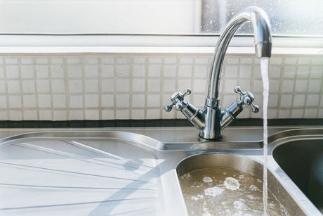 Stainless steel kitchen sink with running water from a chrome dual-handle mixer tap.