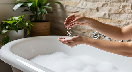 Hands sprinkling bath salts into a warm bubble bath beside a green plant