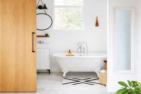 Bright bathroom with freestanding white clawfoot bathtub, sliding wooden door, and minimalist vanity in natural light