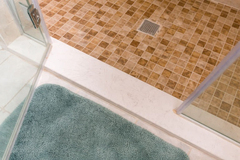 Walk-in shower entrance with mosaic tile floor, stainless steel drain, glass door, and bath mat.