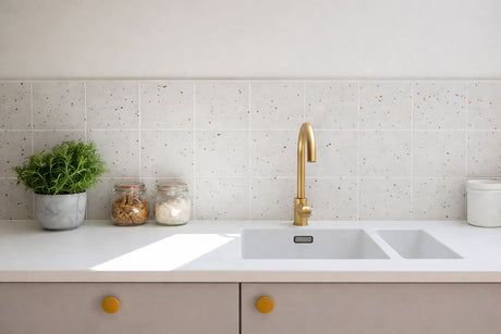 Modern kitchen sink with brushed gold faucet, white countertop, terrazzo tile backsplash, and minimalist decor.