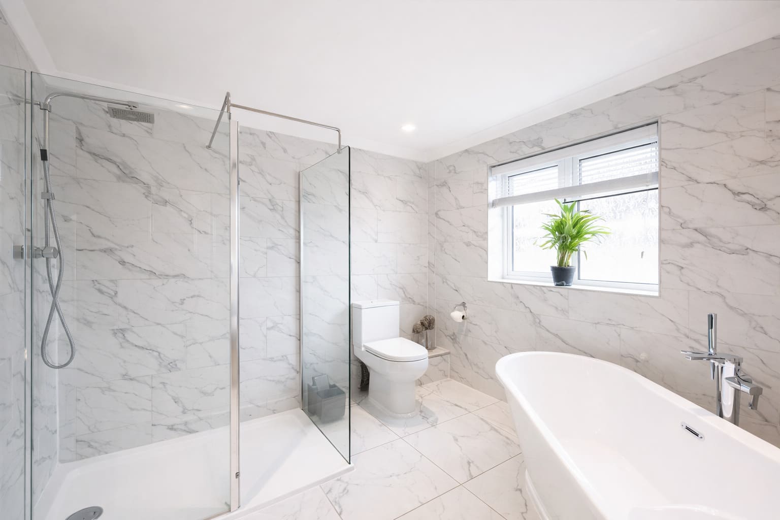 Modern marble bathroom with glass walk-in shower, freestanding bathtub, toilet, and window with indoor plant.