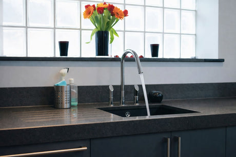 Modern kitchen sink with chrome faucet running water, dark stone benchtop, and fresh flowers by the window.