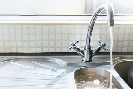 Stainless steel kitchen sink with chrome faucet pouring water into the basin, against a tiled backsplash.