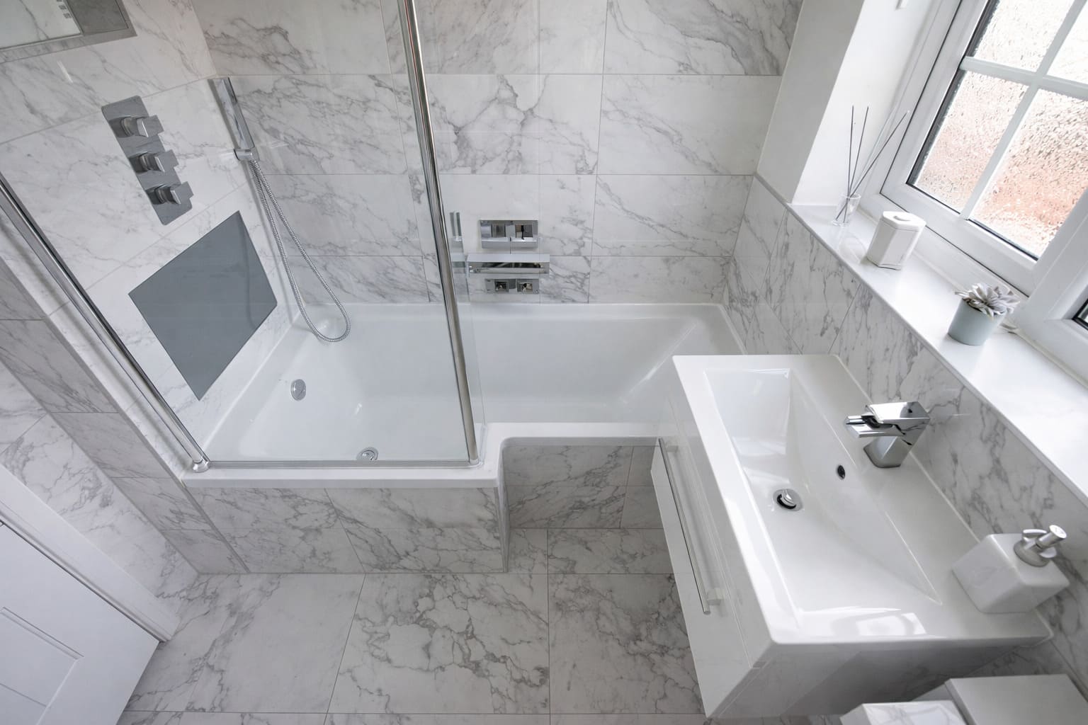 Modern bathroom with marble-look tiles, glass shower over bath, compact vanity basin, and chrome tapware.