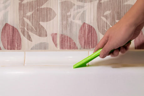 Close-up of hand cleaning grout with green brush between bathtub and decorative tiles