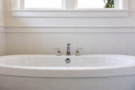 White freestanding bathtub with chrome tapware, beadboard wall paneling, and natural light from window.