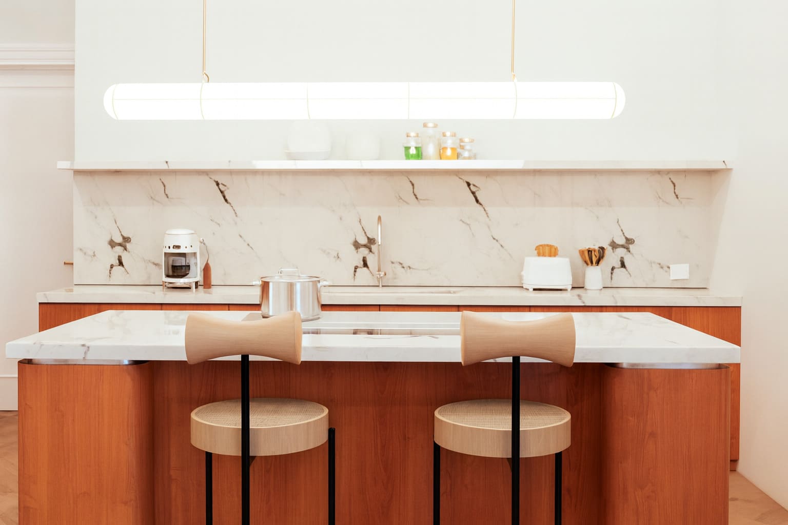 A modern kitchen with a marble island, wooden cabinetry, and two bar stools under a sleek pendant light.