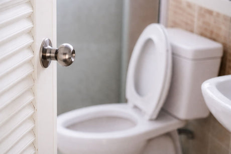 White bathroom toilet with seat open viewed through louvered door, stainless steel doorknob in focus, modern tiled interior in background