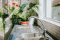 Stainless steel kitchen sink with running water and chrome mixer tap in a bright, plant-filled kitchen.