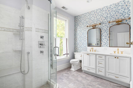 White bathroom with double vanity, glass shower, blue patterned wallpaper, and brass fixtures.