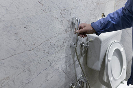 Plumber in blue uniform holding chrome bidet spray beside white toilet and marble tiles
