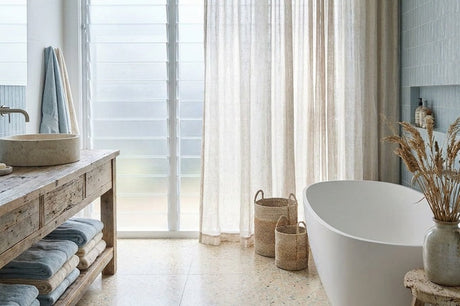 Coastal bathroom with white freestanding bath, rustic timber vanity, sheer linen curtains and woven baskets