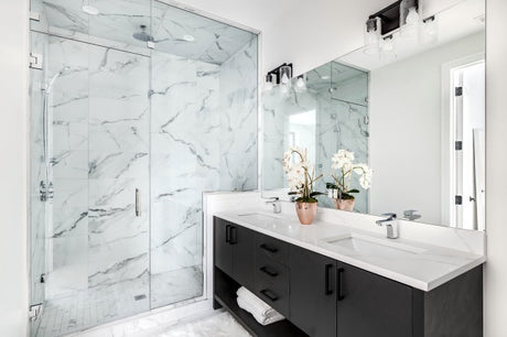 A stylish bathroom featuring a dark timber vanity with a white stone counter top, brass tapware, and marble-lined shower in a modern renovation