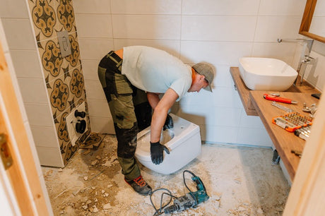 Worker installing a toilet during bathroom renovation with tools and tile debris nearby.