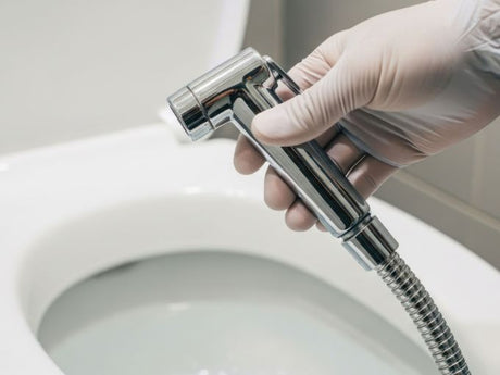 gloved hand holding handheld bidet sprayer next to toilet bowl in bathroom