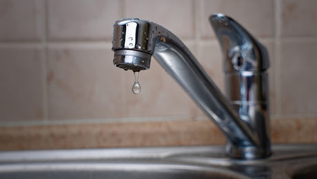 Chrome kitchen tap dripping water over stainless steel sink, beige tiles behind