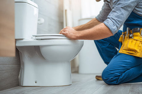 Plumber in blue overalls and tool belt fitting a white toilet seat