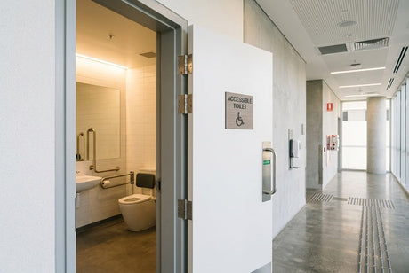 Open accessible toilet door with grab rails, grey hinges, and corridor beyond