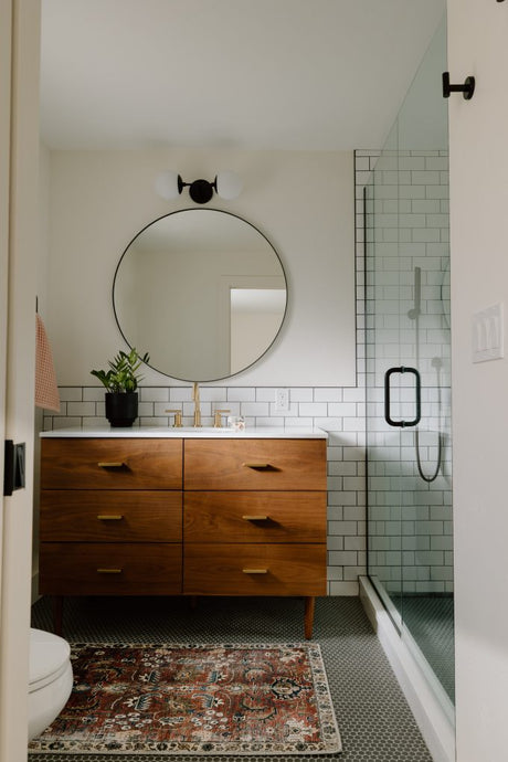 A modern compact bathroom featuring a wooden vanity, round mirror, glass shower screen, and white subway tiles with natural light
