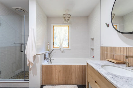 White tile bathroom with wood bathtub panel, glass shower, marble vanity, and round mirror.