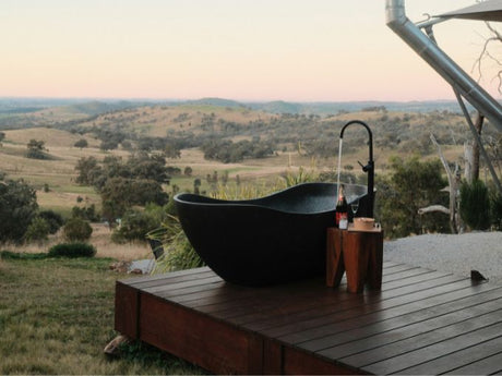 Outdoor freestanding bathtub on wooden deck overlooking countryside landscape with hills and trees at sunset