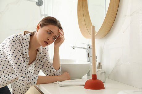 Young woman sitting on bathroom floor looking frustrated while holding a plunger near a blocked toilet