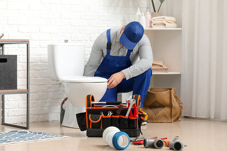Plumber repairing toilet in bathroom with toolbox and plumbing tools on floor.