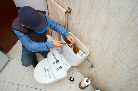 Plumber kneeling beside a toilet repairing the cistern mechanism with plumbing tools on a tiled bathroom floor