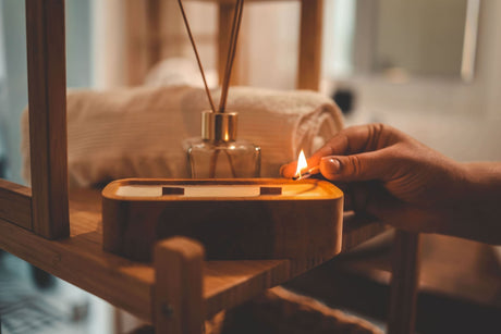 Wooden wick candle being lit on a wooden stool in a dimly lit bathroom with a rolled up towel in the back.