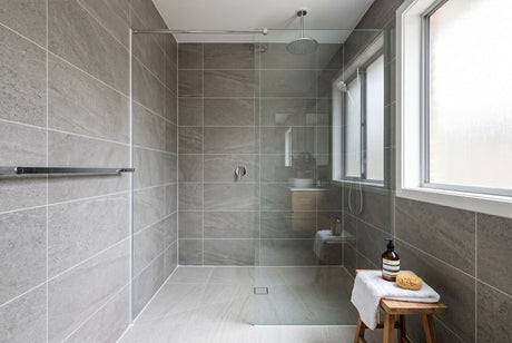 Modern grey stone-tiled shower with glass screen, ceiling rain head and timber stool