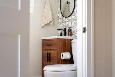 Small bathroom with wood vanity, toilet, round mirror, and floral wallpaper.
