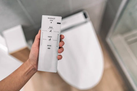 Hand holding a smart bidet washlet remote control with front wash, rear wash, and dryer buttons in a modern bathroom