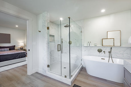 White marble bathroom with glass shower, freestanding tub, wood floor, and bedroom doorway.