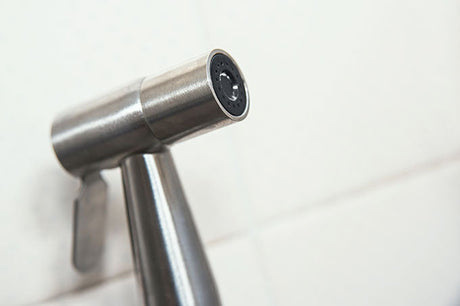 Close-up of a brushed stainless steel bidet spray nozzle against white tiles