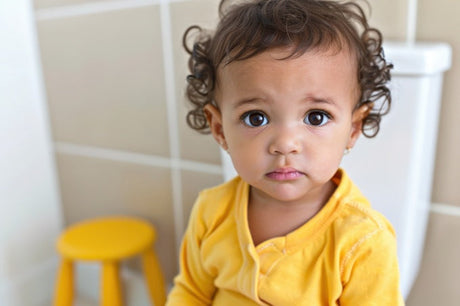 Curly-haired toddler in yellow top sitting on toilet in tiled bathroom