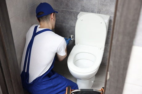 Plumber in work uniform using a spanner to repair a toilet bowl in a white tiled bathroom