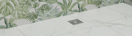 Square stainless floor waste set in white marble tiles with leafy wall backdrop.