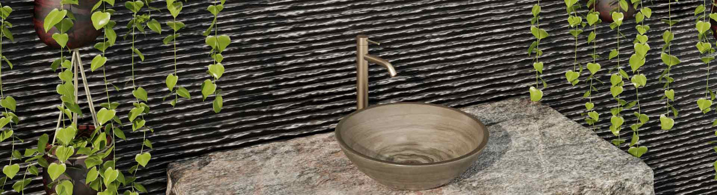 Bronze outdoor basin mixer tap paired with round stone vessel sink, surrounded by hanging plants and textured black wall.