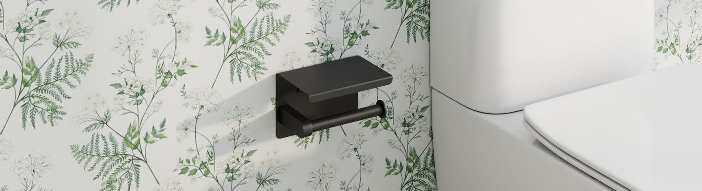 Matte-black toilet paper holder with a small top shelf mounted on a white wall with green fern motifs beside the toilet.